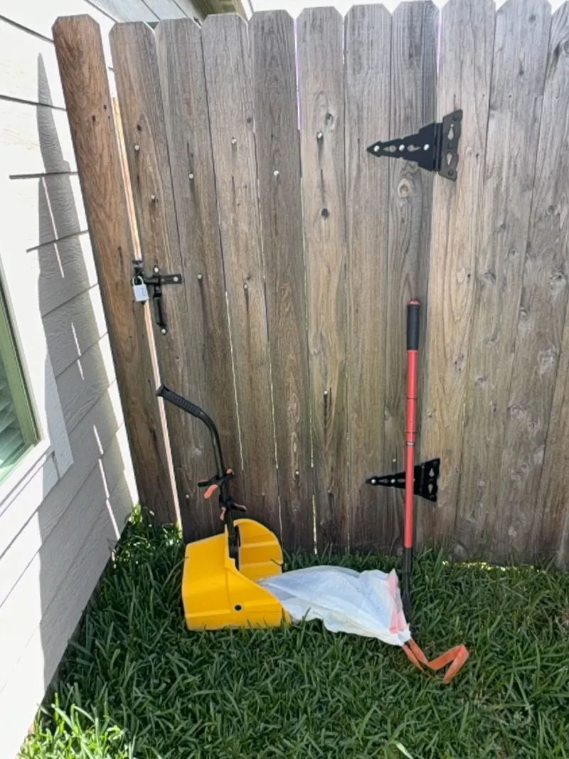 A closed and latched wooden backyard gate after a visit, with the scoop rake, bucket, and bagged waste leaning beside it ready to go on the truck