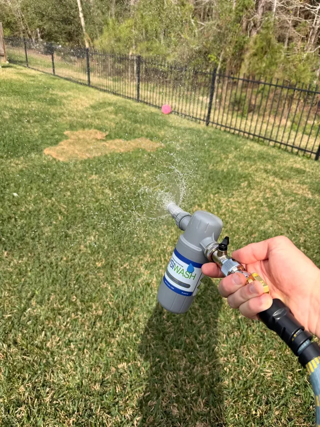 A technician's hand spraying a backyard lawn with a Wysiwash hose-end sanitizer, black metal fence behind